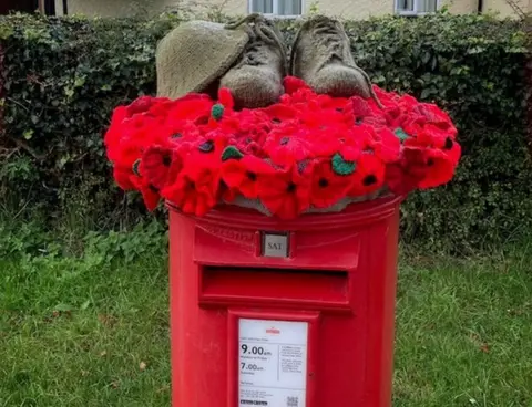 Mark Williamson Red post box on grass with a hedge behind. A garland of poppies lies on top, with knitted soldier's shoes and helmet