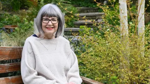 Lorelai is sat in her garden. She is wearing glasses and a cream coloured jumper.