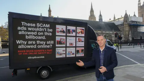 PA Media Martin Lewis poses next to a billboard showing scam advertising