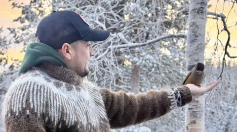 Danny Welch A side view of Danny Welch wearing a thick brown and cream jumper, a blue baseball cap and a green hoody is sticking out from his jumper. His left hand is outstretched and a bird is resting on his hand, eating seeds. Behind him are snow covered trees and a silver birch tree's trunk. 