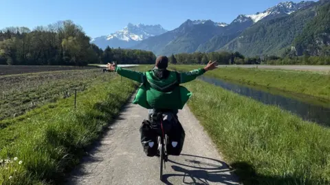 @devonbaistobyronbay A man on a bike facing away from the camera. He is wearing a green coat. His arms are up in the air. He is cycling on a grey path with grass and a river on the right. 