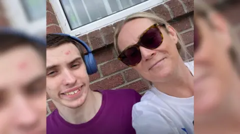 Caroline Campbell A young boy with short dark hair is sitting next to his mum who has blonde hair tied back. He has blue headphones on his head and a purple t-shirt, while she is wearing sunglasses and a white t-shirt.