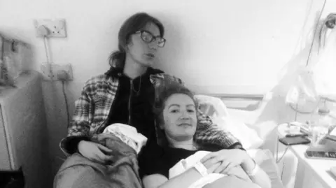 Lowri Denman A black and white photo of Lowri in hospital in September 2015 waiting for scans with her sister. Lowri is lying in bed and her sister had her arm around her. Both have their hair tied back. Lowri's sister is wearing a hoodie.