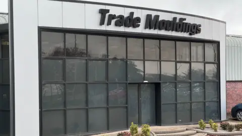 BBC The front of the Trade Mouldings factory in Cookstown.  The cladded building has a large wall of blacked-out windows at its entrance and a large black sign showing the firm's name. 