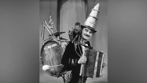Black and white image from 1936 of a man with musical instruments attached to his front and back. 