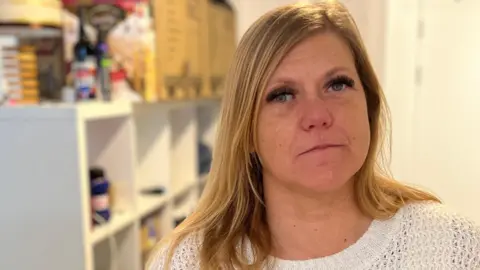 A woman in her late thirties with long light blonde hair wearing a white top and looking at the camera. Behind her are som bags and boxes up on a shelving unit.