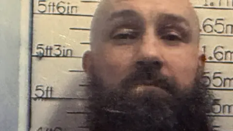 Michael Ross. he has a bald head and a big brown, bushy beard. He is standing in front of a height chart and looking at the camera. 