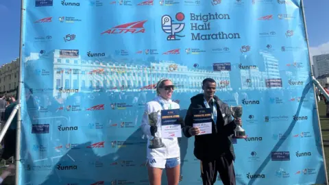 Alison Ferns/BBC The winners of the men's and women's Brighton Half Marathon event stand with their trophies in front of a pale blue plastic sheet with the sponsors' names across it.