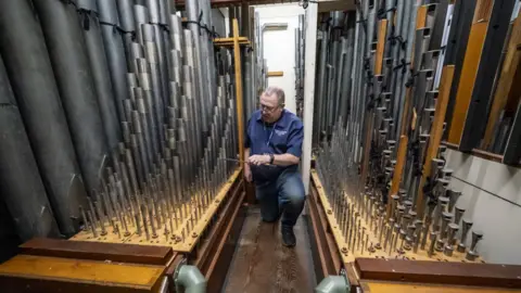 PA/Ben Birchall A man in the organ loft, in between two lines of pipes. He is bending down to fine tune the organ with a reed knife. He is wearing jeans and a blue shirt with a lanyard