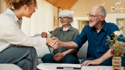 Financial advisor, handshake and old couple with documents for insurance, saving or planning. Retirement, shaking hands and person with senior man, woman and paperwork for mortgage or pension in home - stock photo