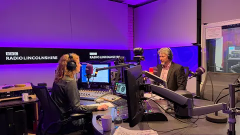 Mike Sheridan is talking to radio presenter Frances Finn. Frances has ginger curly hair and is sat one side of a desk with a microphone, she is looking at Mike Sheridan who is wearing a black suit with a pink shirt. Both are wearing headphones and surrounded by the purple screens of the BBC Radio Lincolnshire studio.