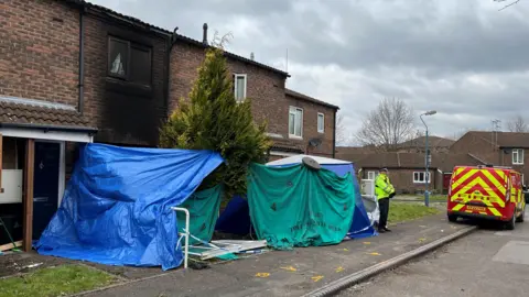 BBC Tents and blue and green material have been set up to obscure the entrance into a terraced house on a residential street. A police officer is standing by the blue tent next to a red fire service van. A wall and window above the tent has been scorched black.