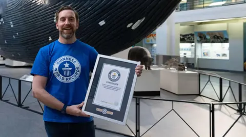 Ben Melham/Guinness World Record Ben Melham in a museum with lots of pictures. He is looking pleased and is holding a tablet with the timing on, as proof he has made the record.