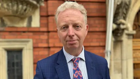 Alex Allinson, who is wearing a blue suit with a shirt and tie and has short grey/blond hair, outside the House of Keys.