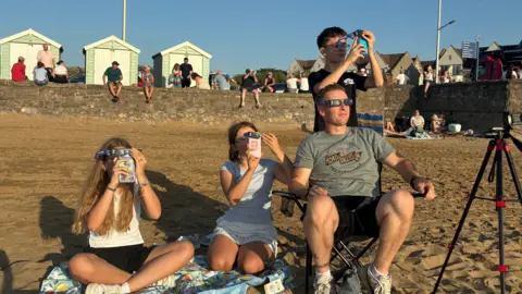 A family of four are sitting on a blanket and a chair while wearing glasses, looking at the eclipse.