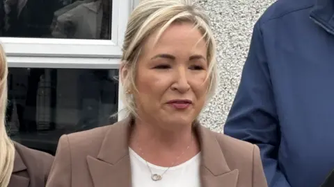 Michelle O'Neill, a woman with blonde hair, wearing a brown jacket and white top,