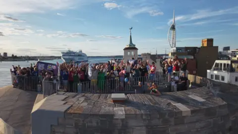 Solent Sky Services A crowd of people standing on the sea wall, with the sea behind including a ferry and Portsmouth's Spinnaker Tower.
The groups are celebrating with their arms in the air, and some are holding banners including one which reads 'WE ARE POMPEY 2029' in white letters on a blue background.