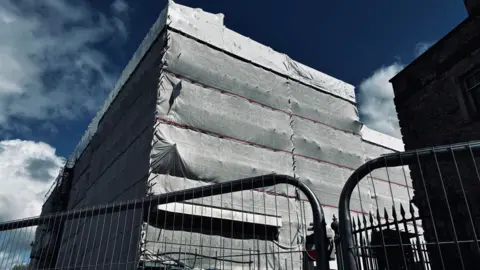 The Evolution Cove apartment block in Plymouth is shown wrapped in white protective sheeting, with metal fencing and barriers placed around the site. The building appears fully encased, with scaffolding visible beneath the covering, suggesting ongoing or emergency safety work. Temporary fencing blocks access at street level, while road barriers and railings mark the edge of the exclusion zone.