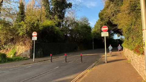 The entrance to Dryden Road with no entry signs in red and white and two people running up the pavement