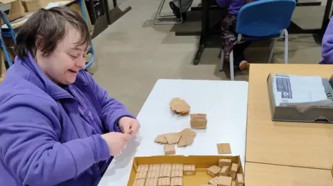 Supported Community Business A person wearing a purple fleece top is piecing together bits of wood at a table. There are other work stations around what appears to be a factory floor. On the table is a box containing small pieces of wood.
