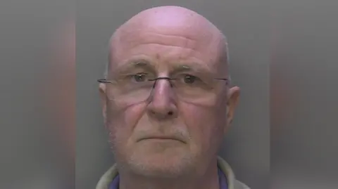 Surrey Police A bald man with glasses and grey stubble stares into the camera for a police mugshot.