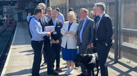 Group of smartly dressed men and women look on as a man in a uniform speaks to them on a train station platform