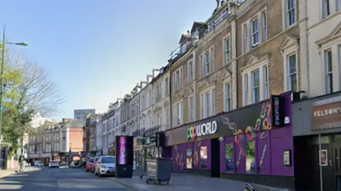 Google Street View image of Old Christchurch Road, a town centre street with cars parked down one side. The street is lined with four-storey Victorian townhouses with the ground floors converted into shops and businesses, including Popworld nightclub in the foreground, which has a purple facade with brightly coloured signage.