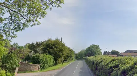 Brock Hill north of Wickford, Essex, a single carriageway with a driveway entry to the right, hedges and trees and a verge, and on the left a long dense hedge with the roofs of farm buildings beyond