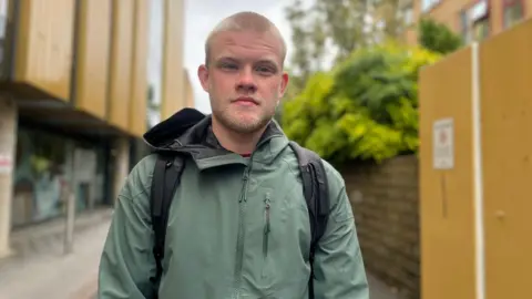Julia Gregory/BBC A man with light blond hair, wearing a light green jacket. He has a backpack and is at a university campus.