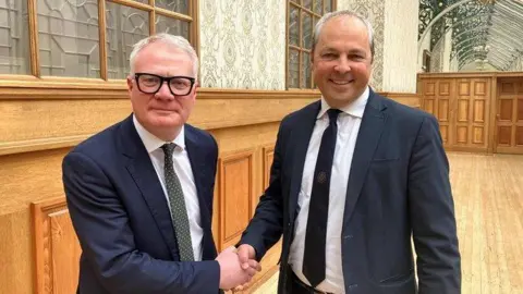 Two men wearing suits shake hands, standing in a wood-panelled room. Both of grey hair and one is wearing thick black glasses.
