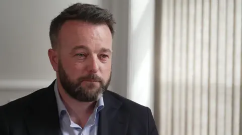 BBC A man with short brown hair and a short brown beard, wears a blue shirt and black suit. He has a small white patch of hair on the bottom right of his beard. There are shut white blinds over his left shoulder and a plane white wall over his right. 
