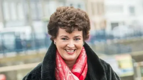 A woman with brown hair wearing a red jumper and a black jacket stands, smiling at the camera. Behind her is a row of houses.