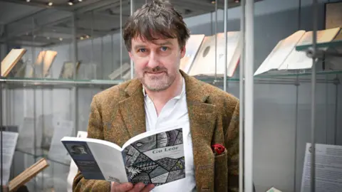 Peter Mackay is standing near display cases containing rare books. He is holding a copy of one his books of poetry.