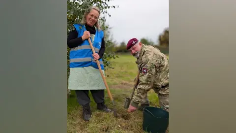 A woman with short brown curly hair, wearing a blue hi-vis vest and a green skirt with black trousers underneath and black shoes, holding a shovel. A man is kneeling next to her wearing army uniform, a maroon beret and holding a shovel. He is kneeling next to a black bucket. He is sprinkling seeds with his left hand. They are both looking into the camera and smiling in a field. 