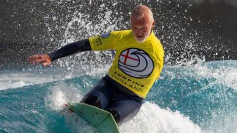 Sarcoma UK Een foto van Stephen Downes op zijn knieën op een surfplank die op een golf rijdt. Hij draagt ​​een geel shirt met de tekst Surfing England over een wetsuit en zijn rechterarm is uitgestrekt voor zijn evenwicht.