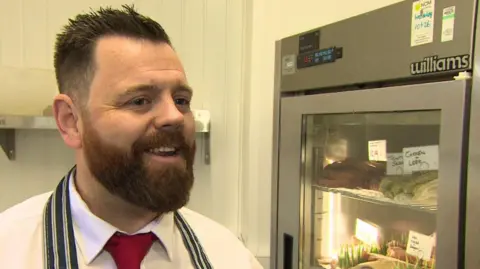 BBC A butcher stands in his shop in front of a fridge full of meat. He has short, spiky hair and a full brown beard and is wearing a white shirt, red tie and blue and white striped apron.