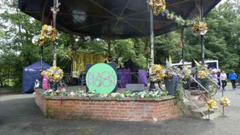 A red brick bandstand has black posts separating it from its black roof. There is a large circular badge which has the words 'Black Moon' written on it. Behind the stand is a navy tent. There are yellow floral arrangements all over the bands railings. As well as a table with a purple table cloth in the centre.