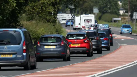 A long line of traffic in the sunshine. The road is single-carriageway with two caravans queued in the distance.