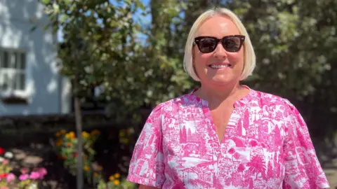 Helen is wearing sunglasses stands in front of a landscaped flower bed and a white building. Trees and greenery fill the background under a clear blue sky.