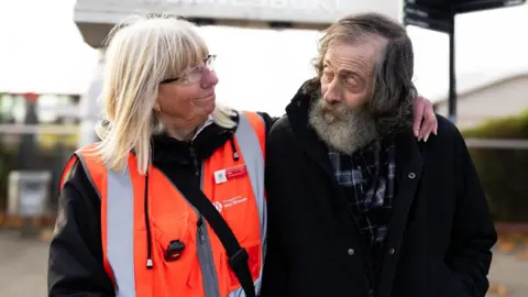 Transport for West Midlands A lady with blonde hair and wearing glasses, with her arm around an older man with grey hair and a beard. They are looking at each other. 