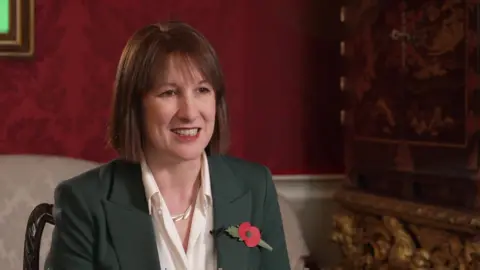 BBC Rachel Reeves speaks from her office sitting on a chair amid the grand surroundings of the Treasury, including gold leaf furniture and red wallpaper. 