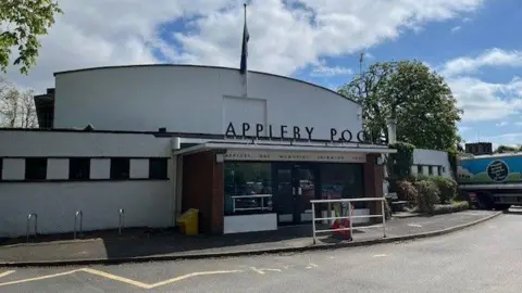 The outside of Appleby Pool. The one-storey white building has the letters spelling out its name above the entrance which has glass doors with a window on each side.