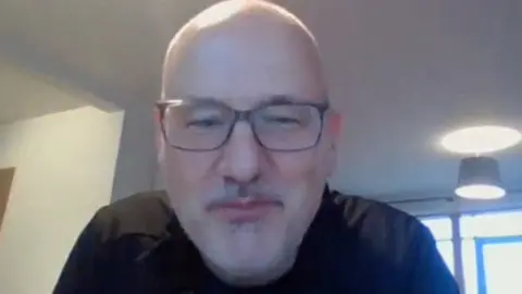 Chris Butler has a shaved head and is wearing glasses and a black T-shirt. This is a still taken during an interview conducted over a video call.