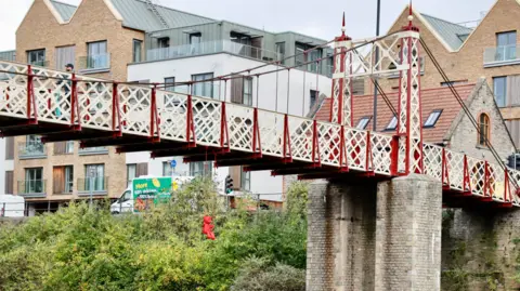 James Colomina A sculpture of a bright red teddy bear on a red swing can be seen hanging from the bridge. The red and white bridge can be seen in the picture, as well as buildings and the road behind it. 