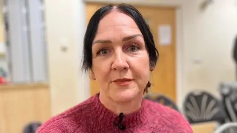 Kate Bradbrook/BBC Maggie Mahon is sat in the middle of the image. She has black hair in a ponytail and is wearing a pink jumper. 