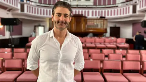 Oliver Tompsett is smiling as he stands in front of rows of seating in a performance space. He has short brown hair and is wearing a white shirt.