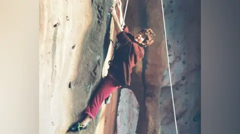 Kendal Wall A woman is climbing on an indoor wall. She is wearing a harness and is holding on the side of the wall which has brown-coloured holds. She has short, brown hair and is wearing maroon stretchy trousers and a baggy, brown jumper. The image appears to be old.