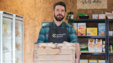 A man in a community grocery apron holds a wooden box with bread in it. He has dark hair, a beard and wears a green and blue checked shirt. Groceries are stacked on shelves behind him.