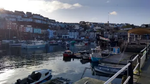 A picture of the harbourside in Brixham. The water in the harbour can be seen with many empty boats on top of the water.