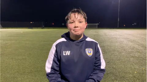 Levi looking at the camera wearing his navy club top. He has a long brown fringe and is standing on the football pitch with a goal a long way behind him.